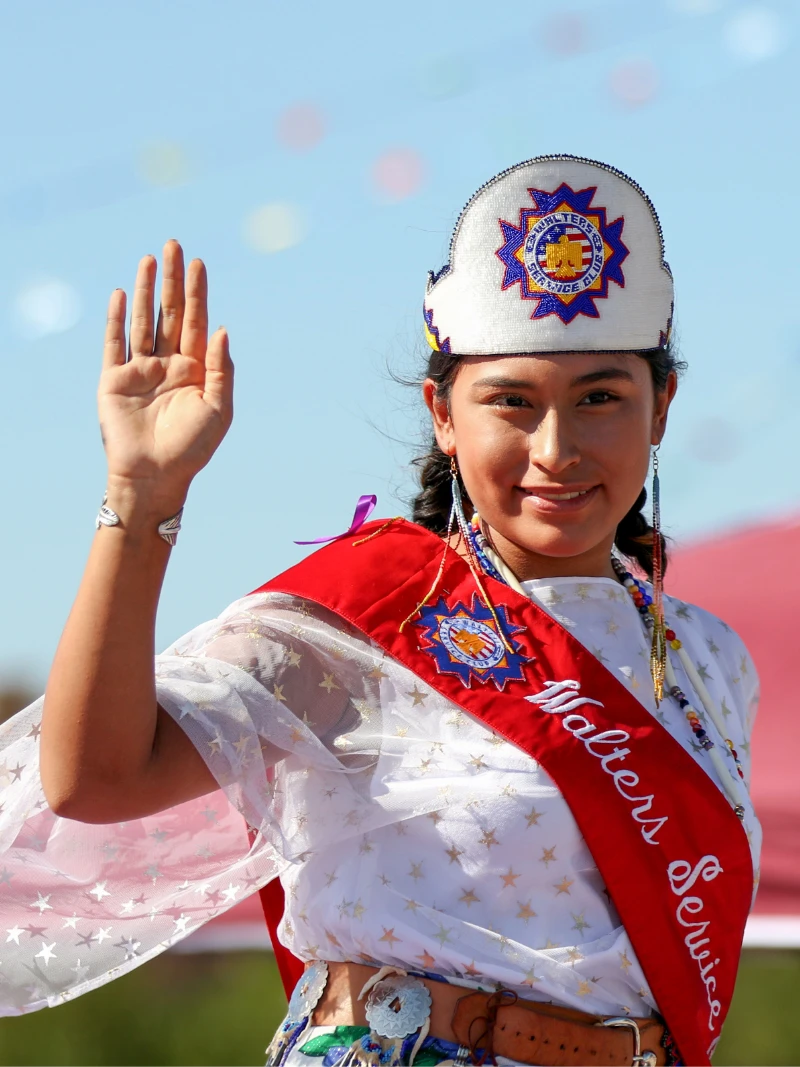 Young woman in traditional attire and sash raises her right hand in greeting, representing the Comanche Nation Tʉitua Foundation. She wears a decorative hat and stands outdoors with a blurred background, proudly Preserving Comanche Heritage.