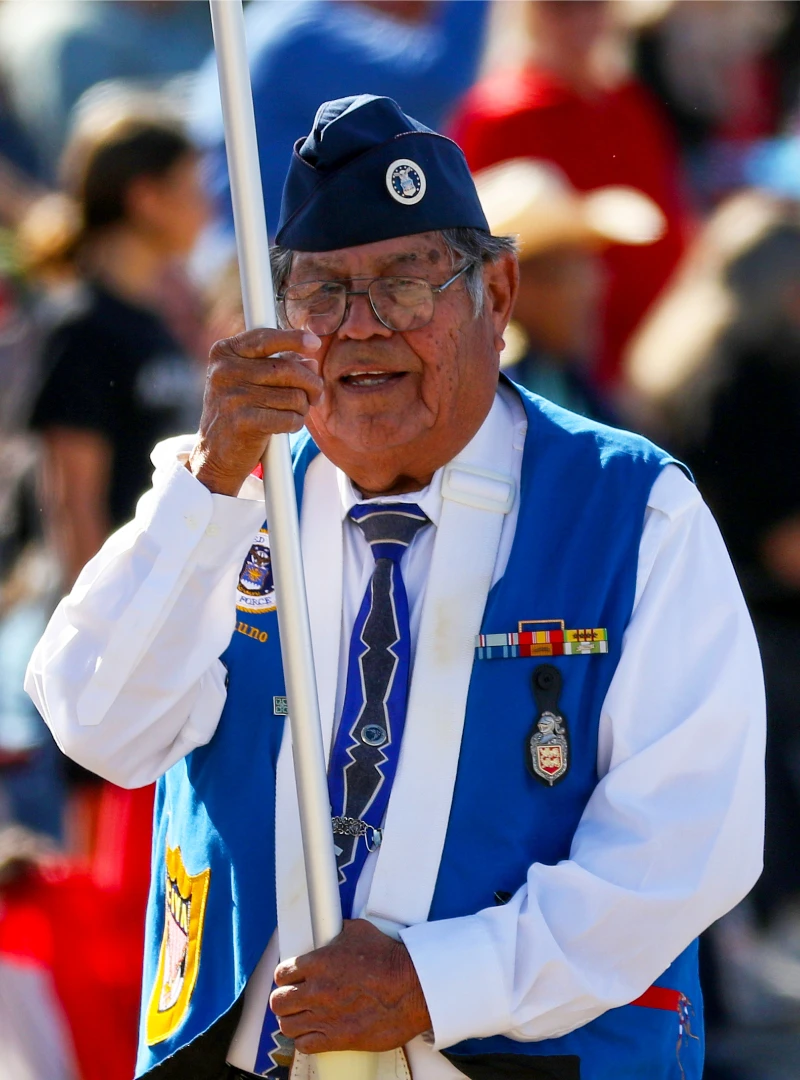 Older man in a blue uniform vest and hat holds a flagpole during a public event, wearing medals, a tie, and glasses, with people blurred in the background.