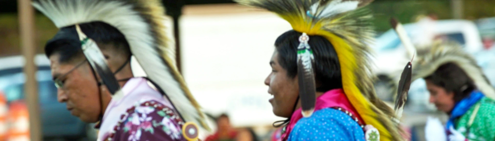 Three people in traditional regalia with feathered headdresses participate in an outdoor event, proudly preserving Comanche heritage and advancing Comanche culture.