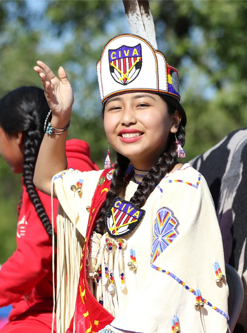 Young person in traditional attire with beadwork, braids, and feathered headdress, smiling and waving outdoors during an event supported by the Comanche Nation Tʉitua Foundation, dedicated to Preserving Comanche Heritage.