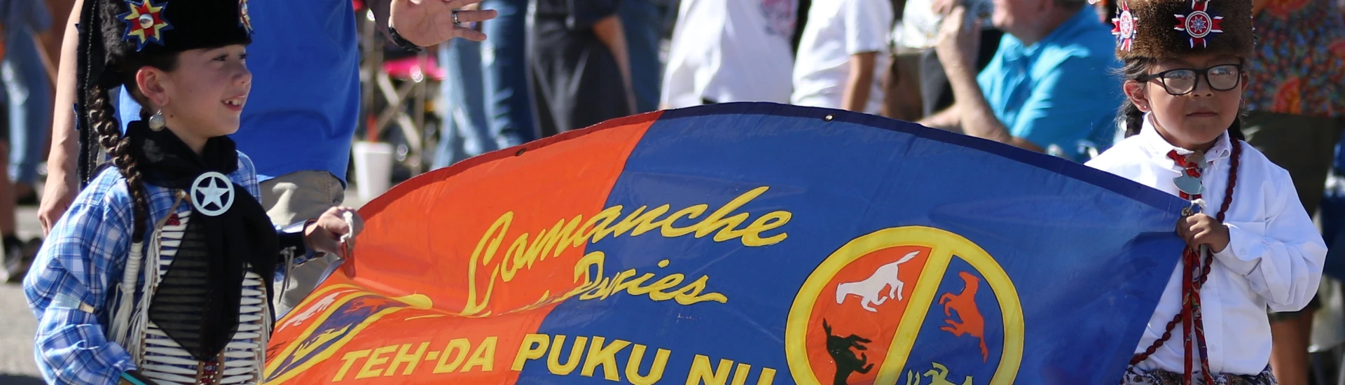 Two children in traditional attire hold a colorful banner at an outdoor event.
