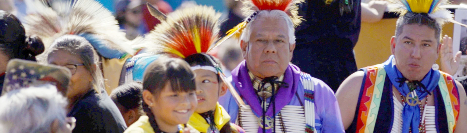 A group of people, including children and adults, wear traditional regalia and feathered headdresses at an outdoor gathering or event, Preserving Comanche Heritage as part of the vibrant activities organized by the Comanche Nation Tʉitua Foundation.