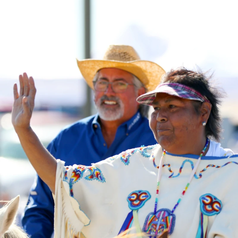 Two people outdoors, one in a straw hat and blue shirt smiling, the other in a decorated garment and visor waving, both participating in a Comanche Nation cultural event that celebrates and advances Comanche culture.