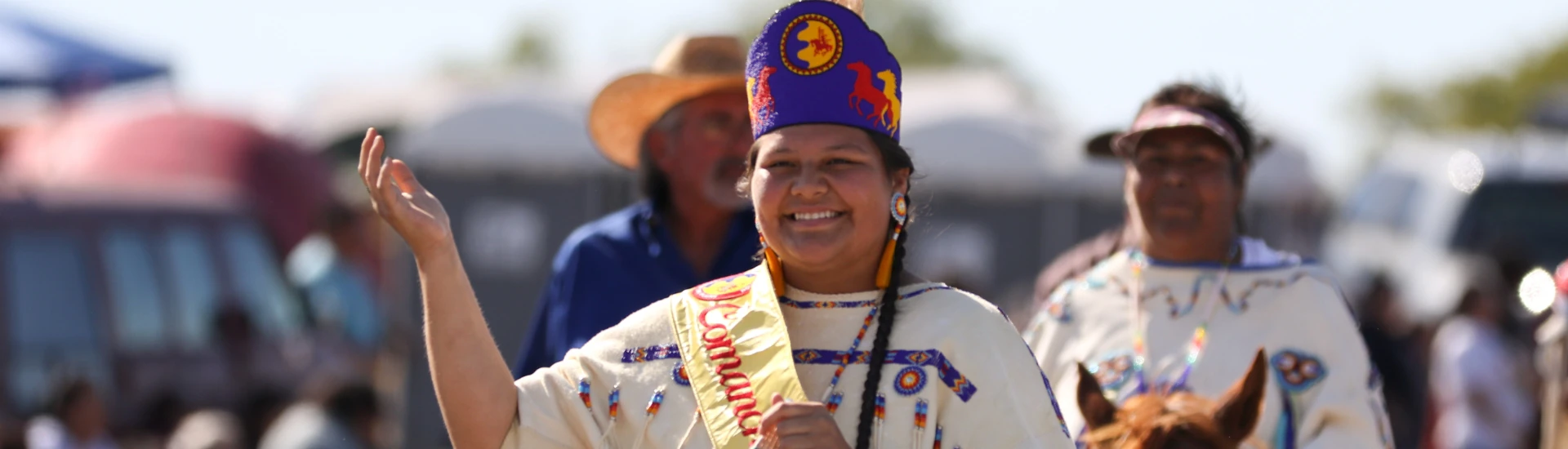 A woman in traditional dress and headdress, representing the Comanche Nation, waves and smiles during an outdoor event.