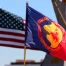 A blue and red flag with the words "Comanche Nation" and “Lords of the Plains” and an image of a horseman, representing the Comanche Nation, is shown next to a partially visible American flag against a blue sky.