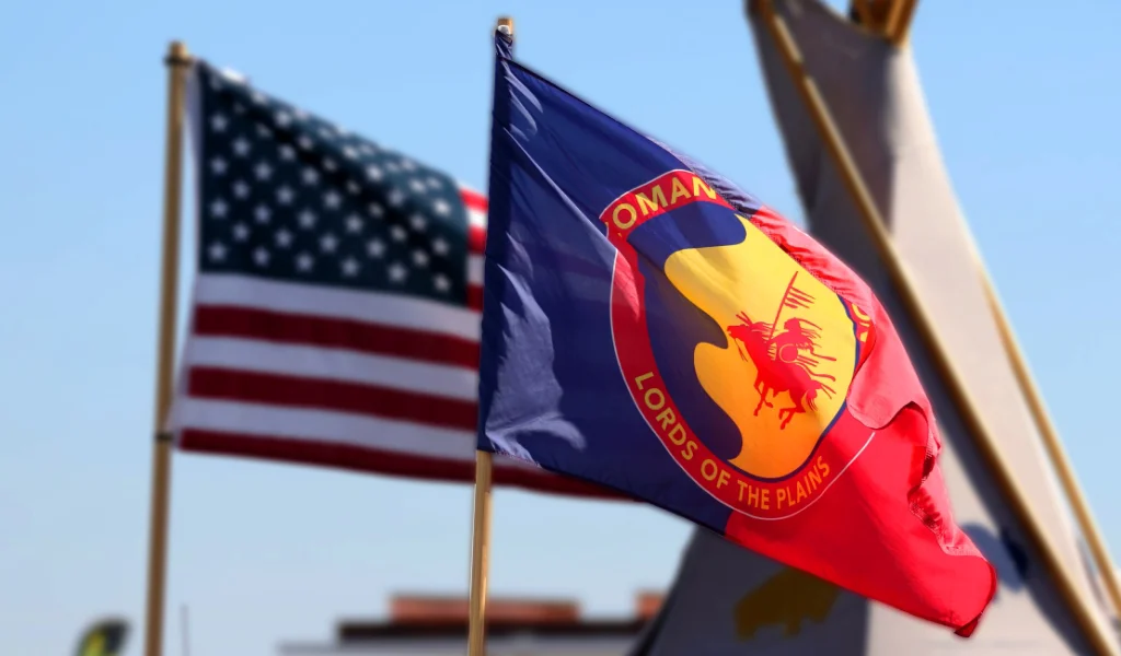 A blue and red flag with the words "Comanche Nation" and “Lords of the Plains” and an image of a horseman, representing the Comanche Nation, is shown next to a partially visible American flag against a blue sky.
